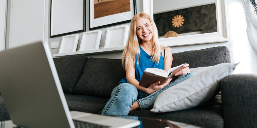 smiling woman with laptop on table reading book on sofa